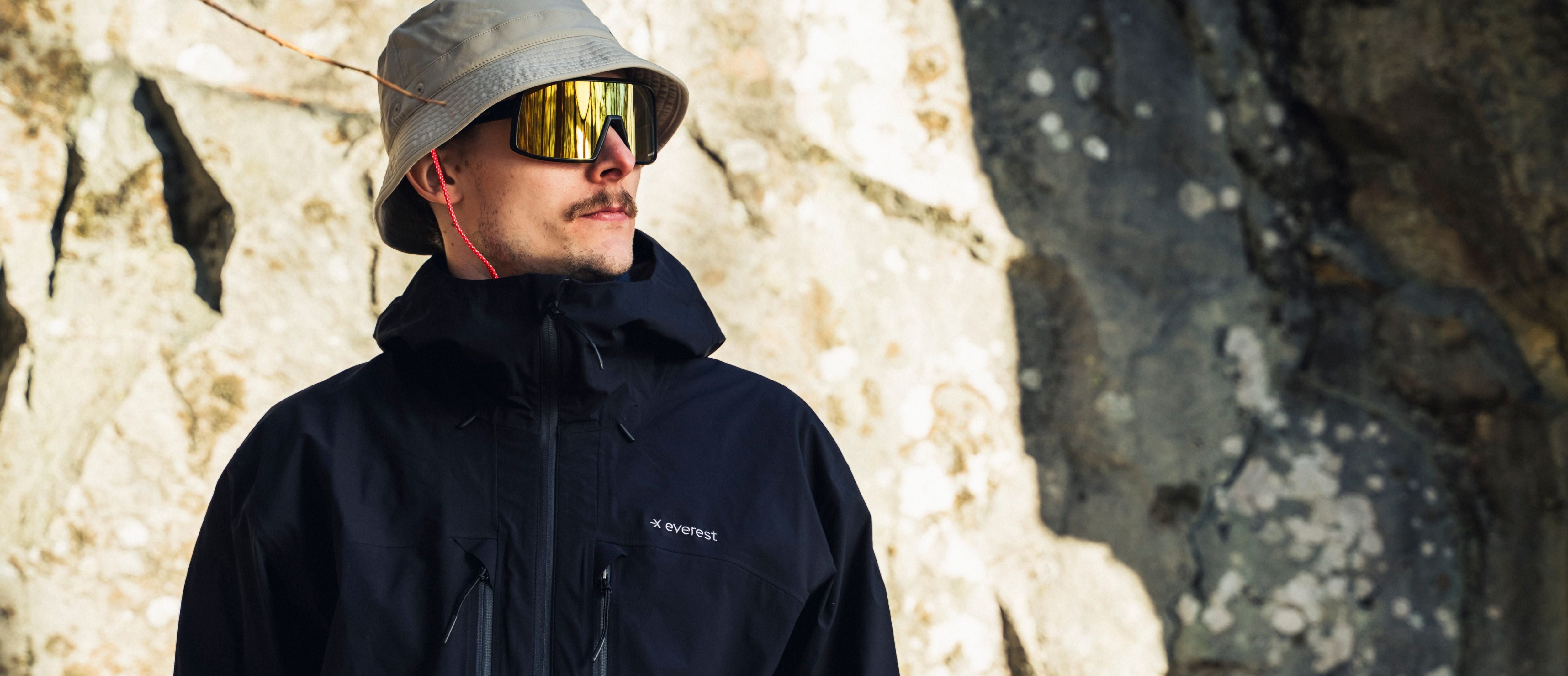 Man wearing a black jacket and gray bucket hat with reflective sunglasses, standing against a rocky background.