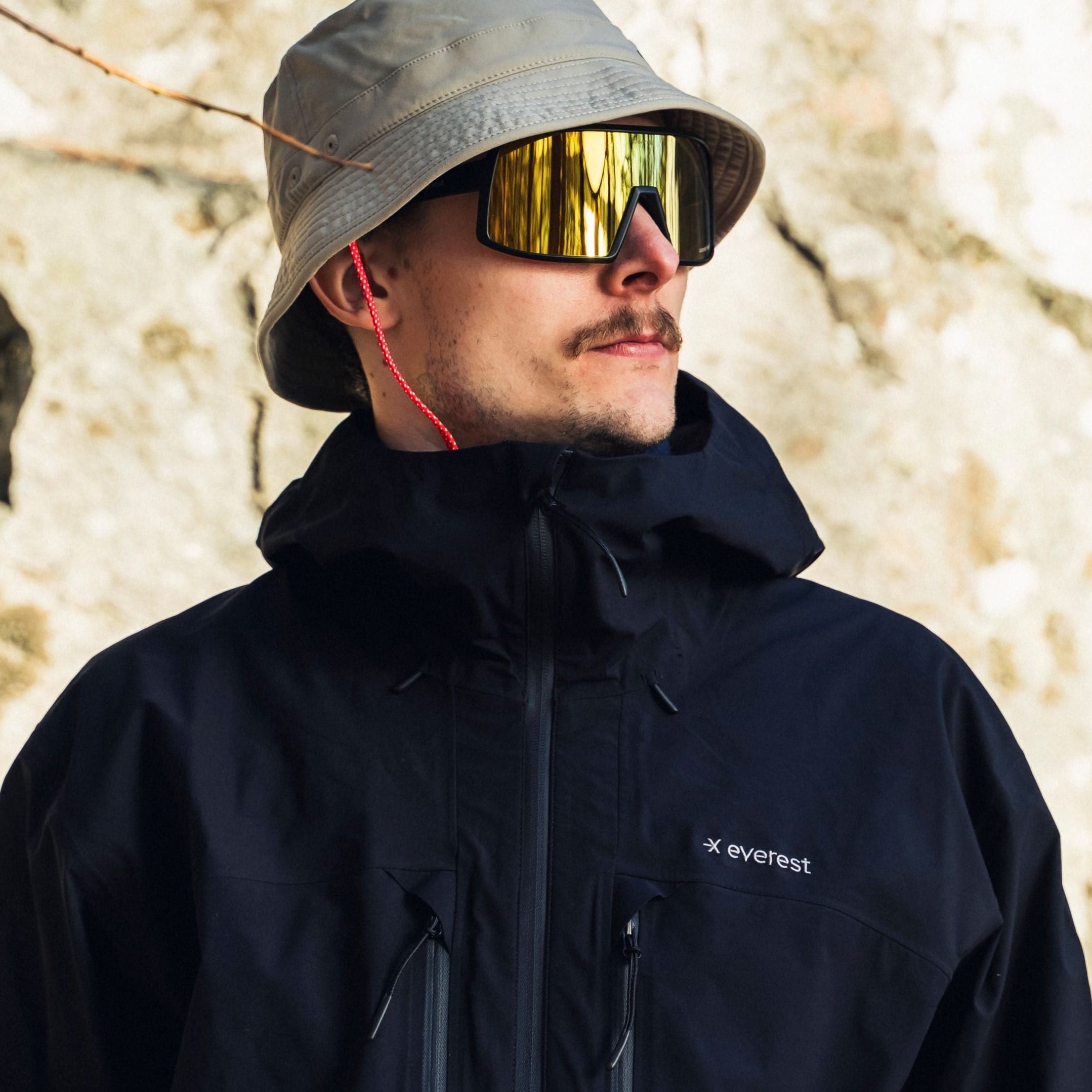 Man wearing a black jacket and gray bucket hat with reflective sunglasses, standing against a rocky background.